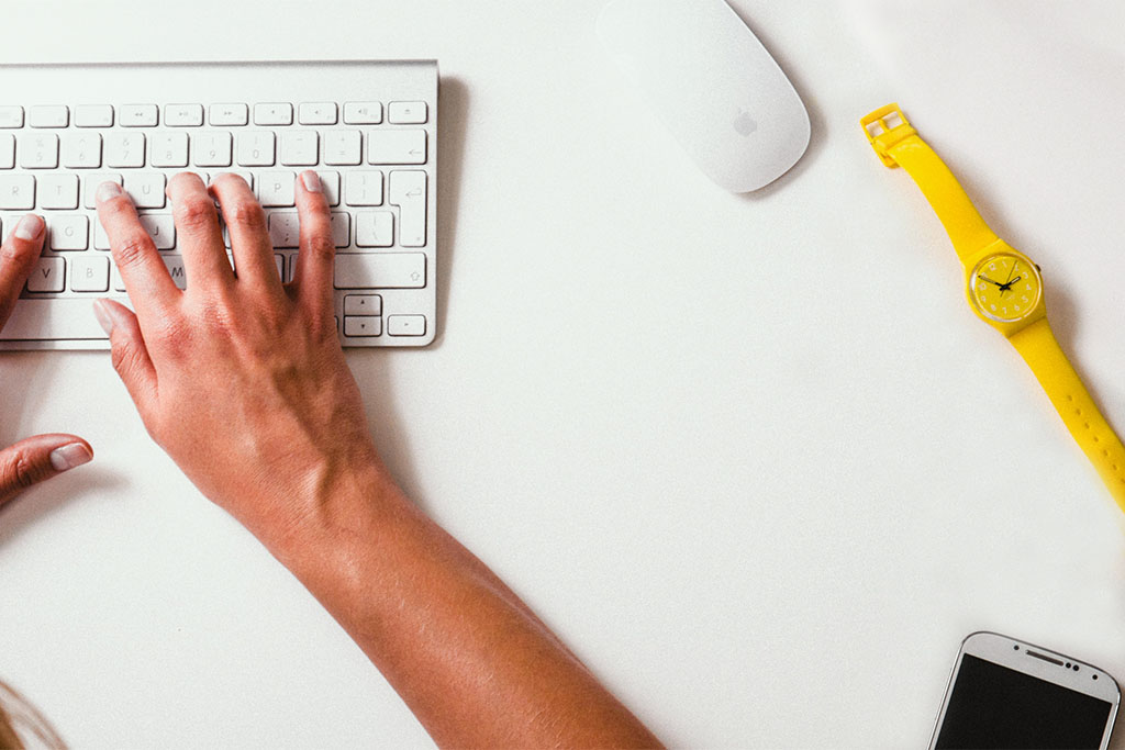 Two hands on a computer keyboard next to a watch, a mouse, and a mobile phone.